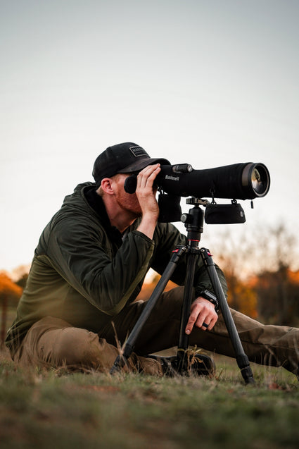 A person wearing a dark cap and green jacket sits on grass, looking through a large spotting scope mounted on a tripod, with a blurred outdoor background.