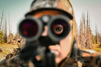 A person in camouflage clothing looks through the scope of a rifle in a dry, barren forest. The focus is on their eye, visible behind the scope, with blurred trees and landscape in the background.