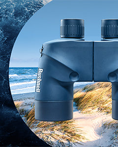 Blue binoculars with the Bushnell logo, positioned in front of a scenic coastal landscape featuring sandy dunes, grass, and ocean waves under a partly cloudy sky.