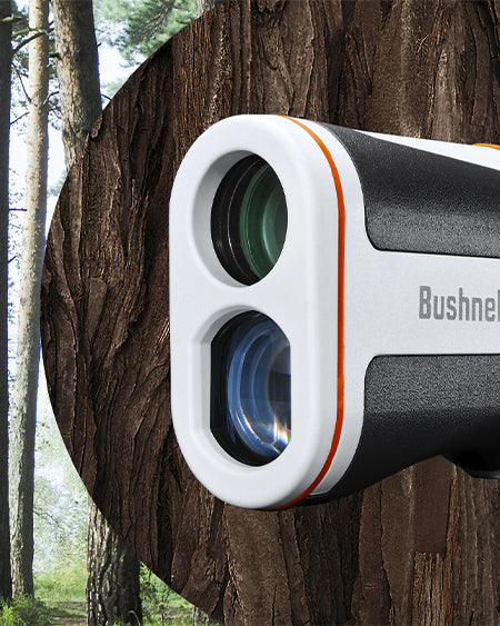 Close-up of a Bushnell rangefinder with white and black casing and orange accents, set against a background of tree trunks and a forest scene.