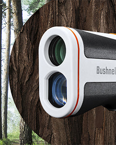 Close-up of a Bushnell rangefinder with a white and black body and two lenses, set against the textured background of a tree trunk and tall trees in a forest.