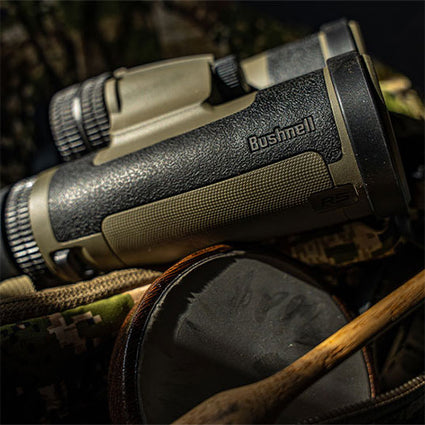 Close-up of a pair of black and olive green Bushnell binoculars resting on camouflage fabric, with a wooden stick and a circular object partially visible in the foreground.