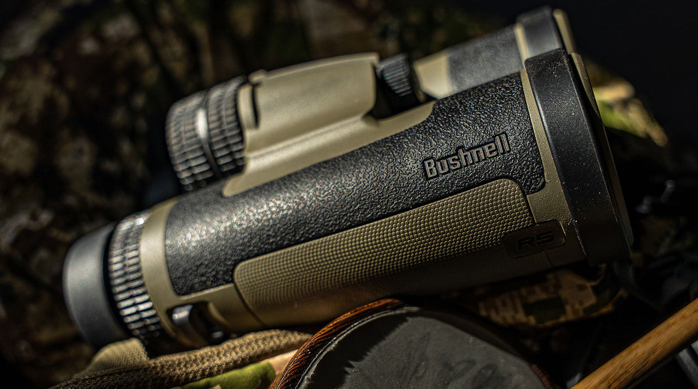 Close-up of a pair of Bushnell binoculars with textured black and olive green grips, resting on camouflage fabric and outdoor gear in natural light.