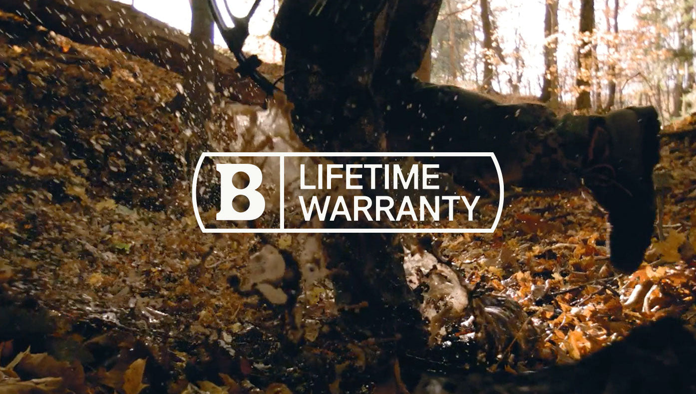A person in camouflage gear runs through a muddy, leaf-covered forest floor, splashing water and mud. Overlay text reads B LIFETIME WARRANTY in a bold white font.