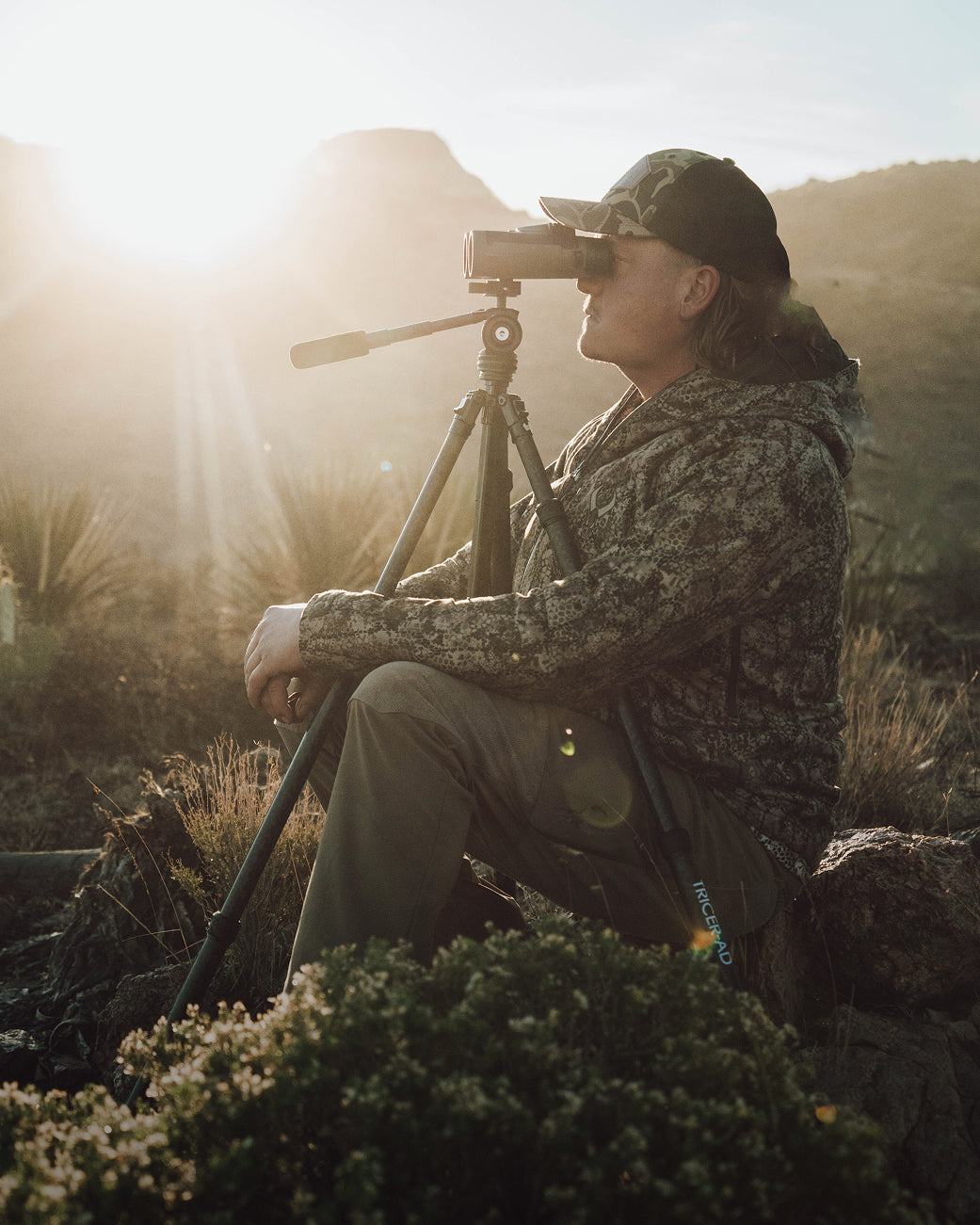 A person in camouflage clothing and a cap sits outdoors on a log, looking through binoculars mounted on a tripod, with sunlight and mountains in the background.
