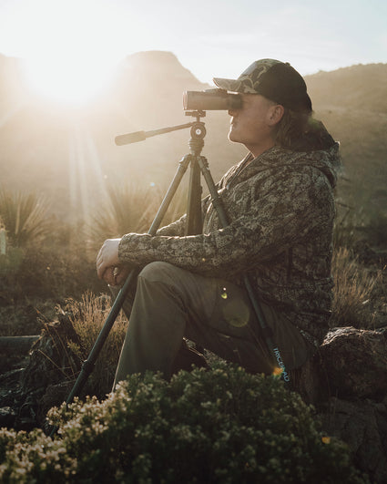 A person in camouflage clothing and a cap sits outdoors on a log, looking through binoculars mounted on a tripod, with sunlight and mountains in the background.