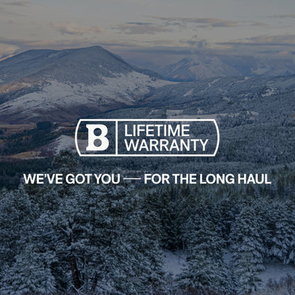 Snow-covered mountains under a cloudy sky with a forest in the foreground. Text in the center reads: “B Lifetime Warranty. We’ve got you — for the long haul.”.