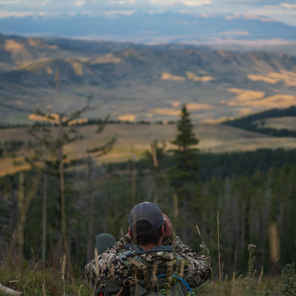 Person in camouflage sitting on a hilltop with a scenic view of mountains and valleys.
