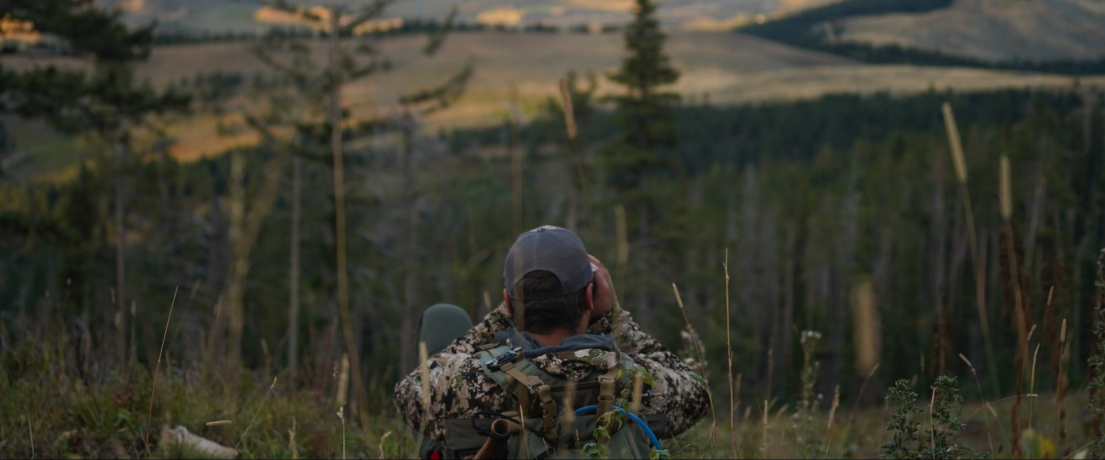 Person in camouflage clothing sitting on a hill with a scenic view of trees and mountains.