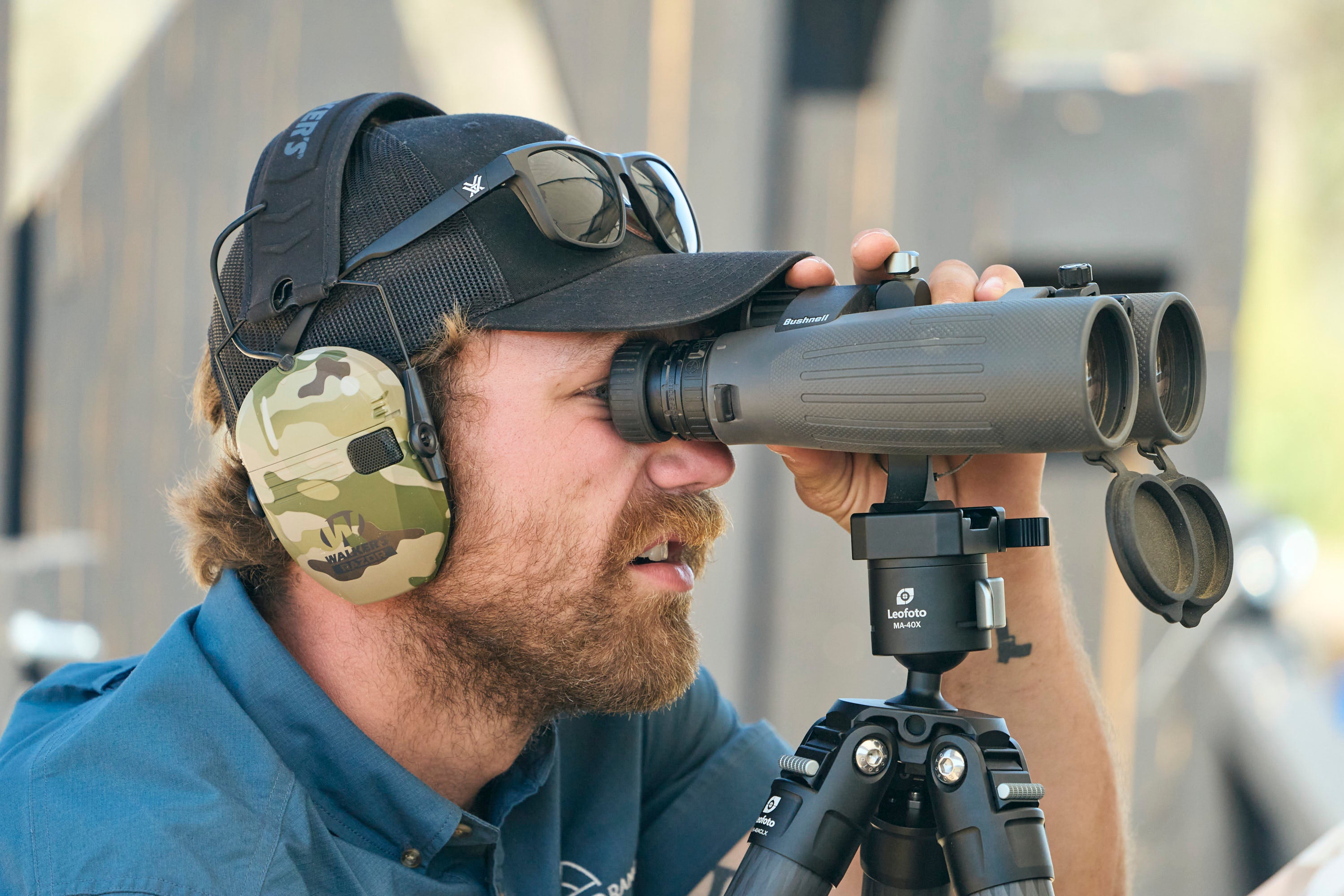 A man wearing a cap, sunglasses, and camouflage earmuffs looks through large binoculars mounted on a tripod, outdoors in daylight.