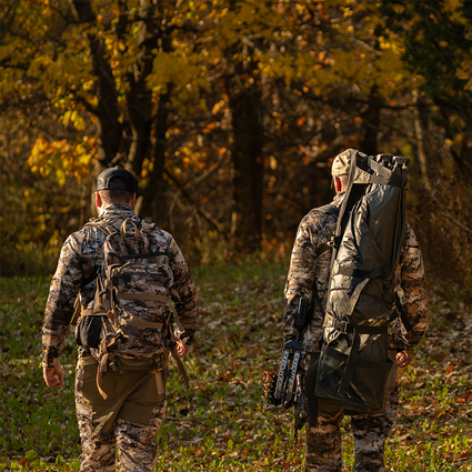 Two hunters in camouflage gear walking through a forest during autumn.