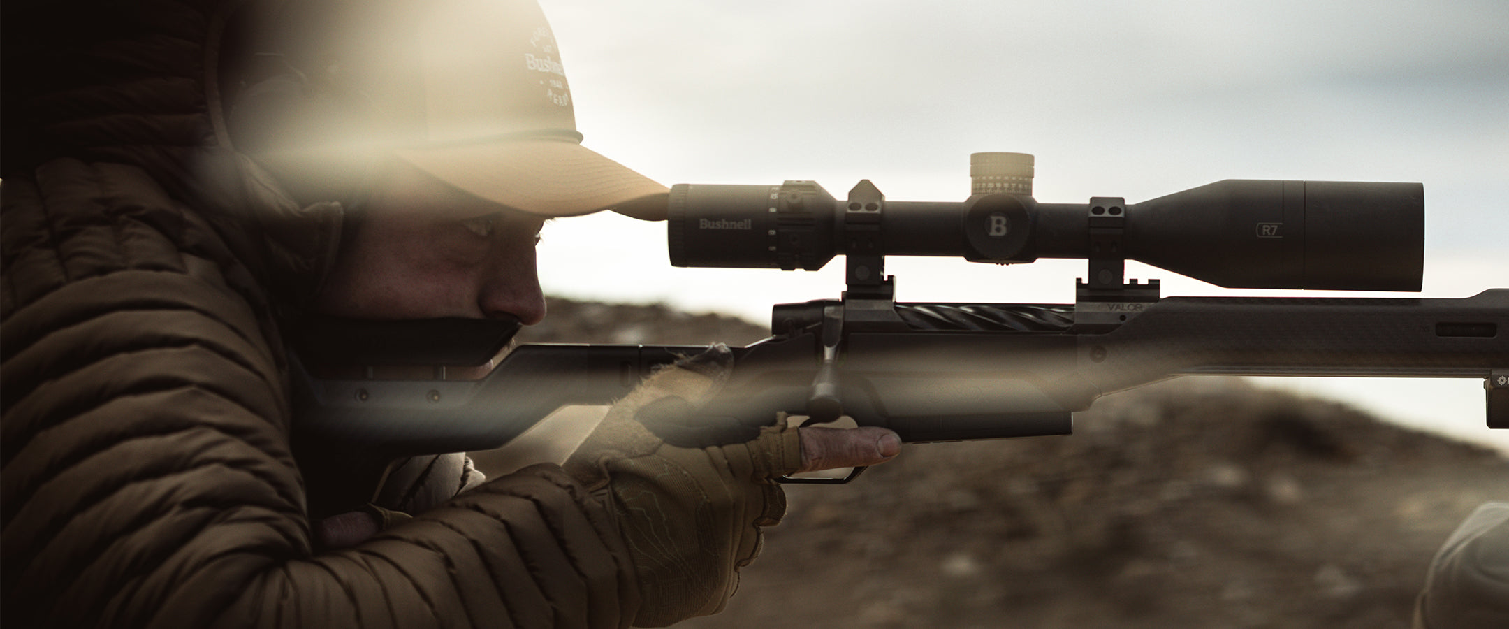 A close-up action shot of a person in a crouched, camouflaged pose aiming a long rifle with a mounted scope. The scene is heavily blurred and tinted in warm, muted earth tones, with bright lens flare on the left.