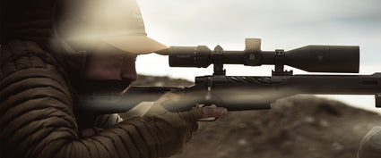A close-up action shot of a person in a crouched, camouflaged pose aiming a long rifle with a mounted scope. The scene is heavily blurred and tinted in warm, muted earth tones, with bright lens flare on the left.
