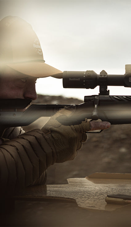 A close-up action shot of a person in a cap and gloves aiming a rifle with a scope in an outdoor setting, with a faint double-exposure overlay of the rifle and scope.