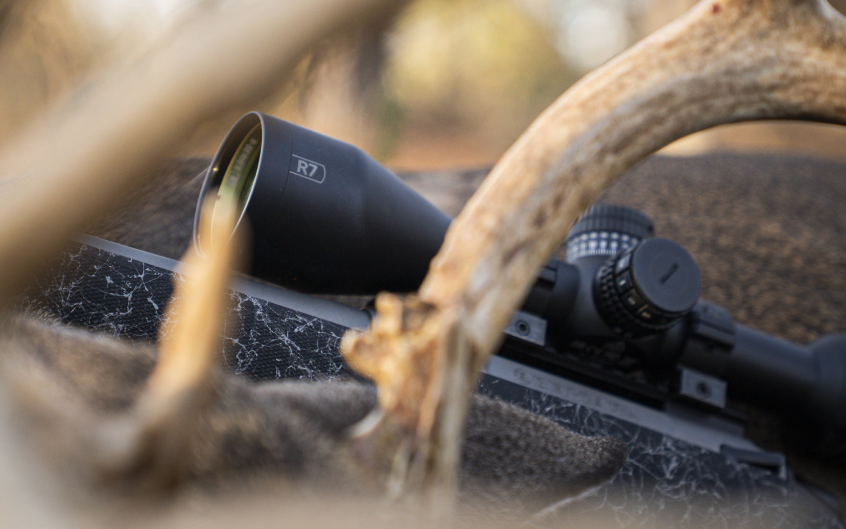 A close-up outdoor shot of a black rifle scope mounted on a camo-patterned rifle, with deer antlers in the foreground and a blurred natural background.