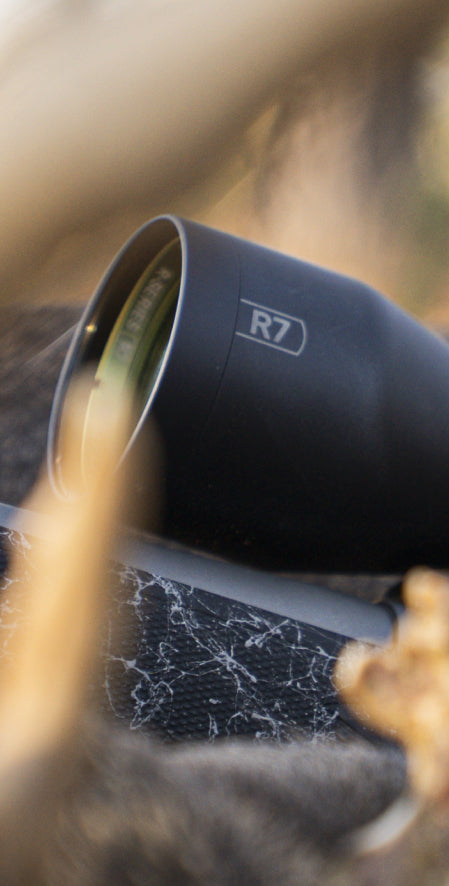 Close-up of a black R7 scope or camera lens mounted on a rifle, with blurred natural background and foreground foliage.