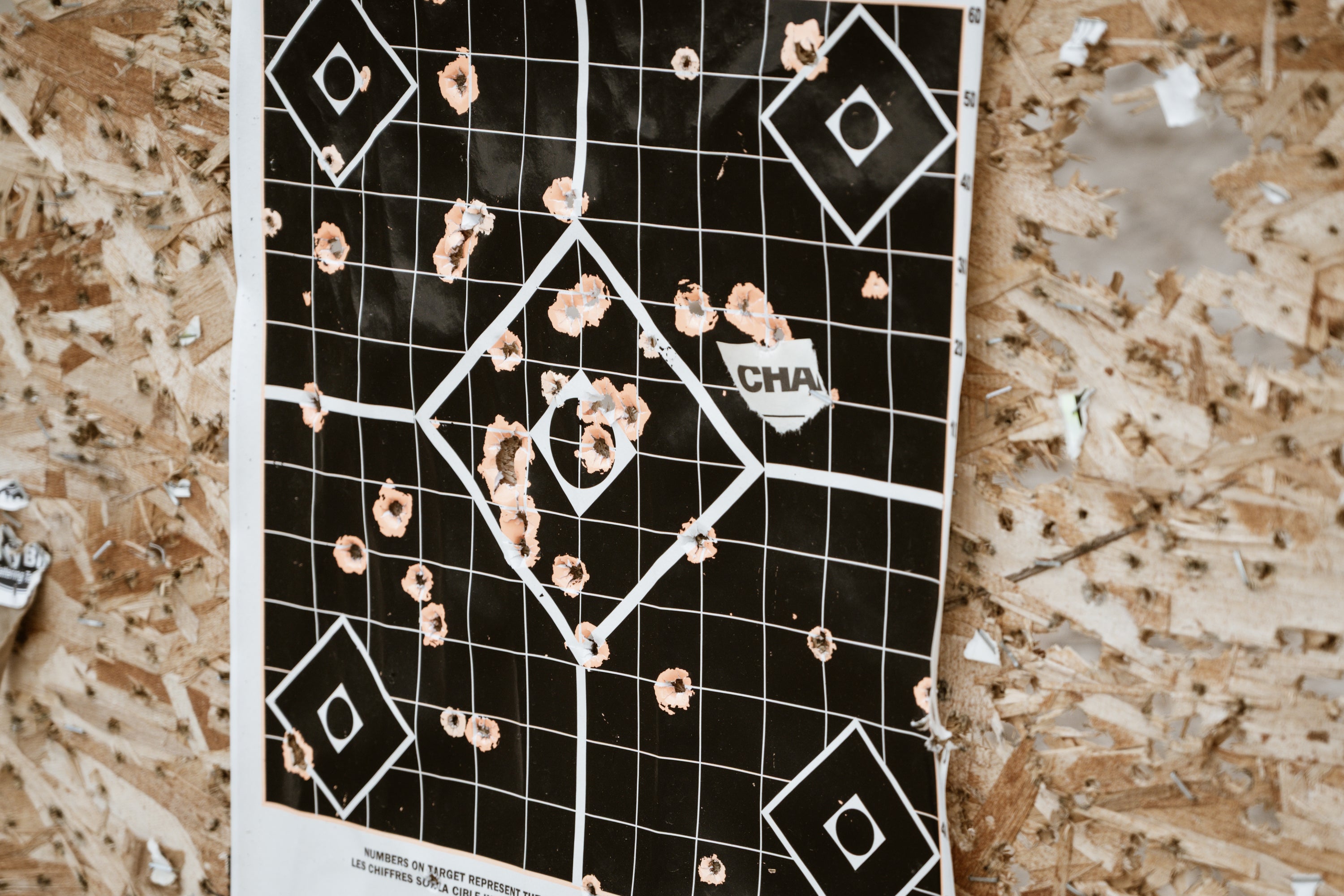 A paper shooting target with multiple bullet holes clustered around the center, attached to a plywood surface. The target features a black grid pattern with white lines and diamond shapes.