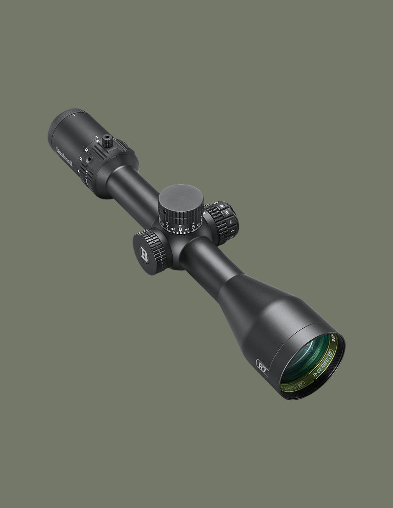 A black tactical rifle scope with adjustment knobs and a green-coated objective lens, shown at an angle against a solid dark olive-gray background.