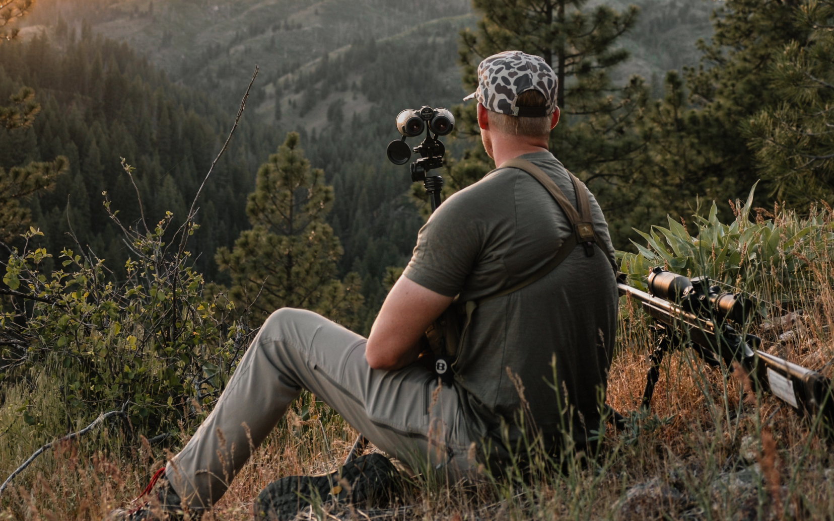 A man in outdoor hunting attire sits on a grassy hillside in a mountain forest, using a spotting scope. A rifle rests on a tripod beside him.