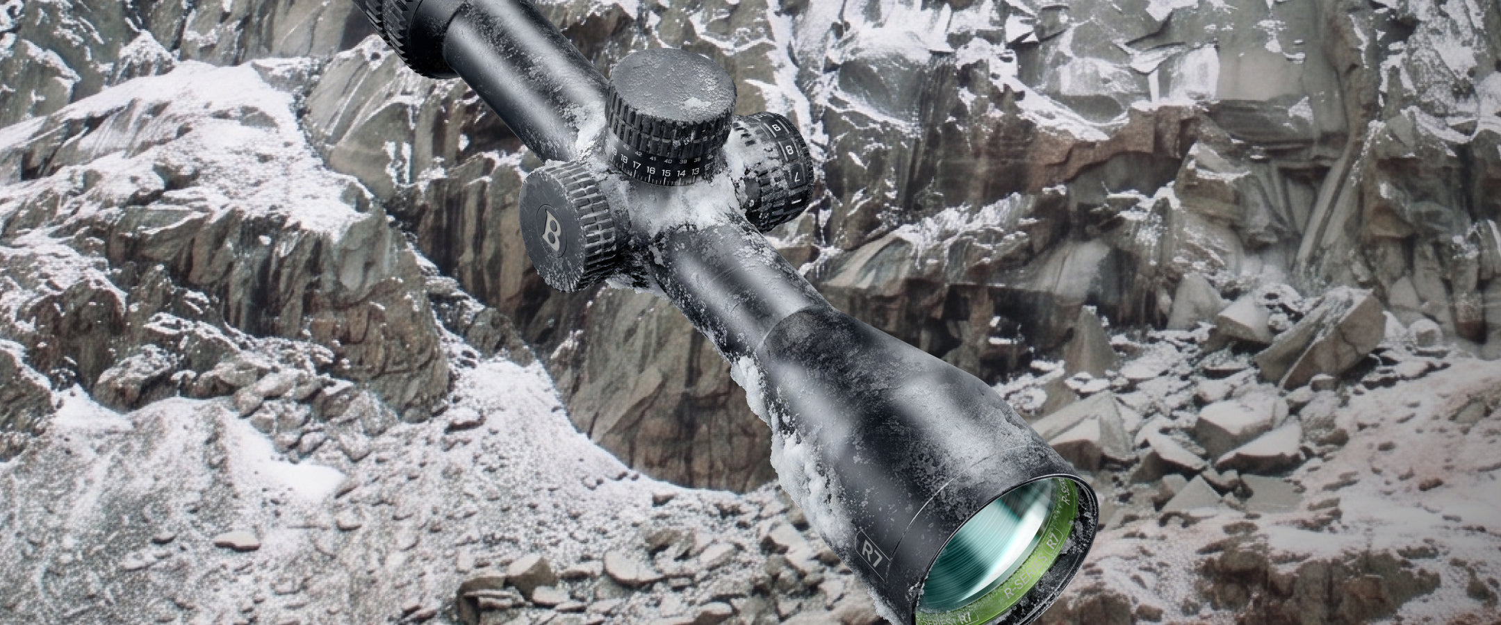 A black rifle scope with a green-tinted objective lens, shown mounted on a rifle and covered in white snow, set against a rocky, snowy outdoor mountain background.