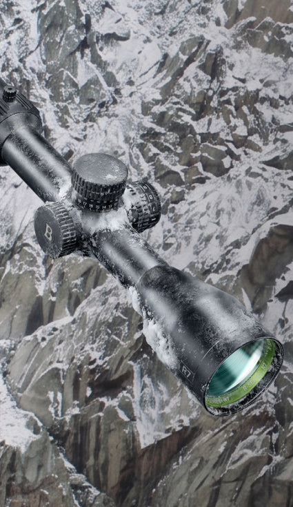 A close-up of a black rifle scope with a green-tinted objective lens, shown against a snowy mountain background.