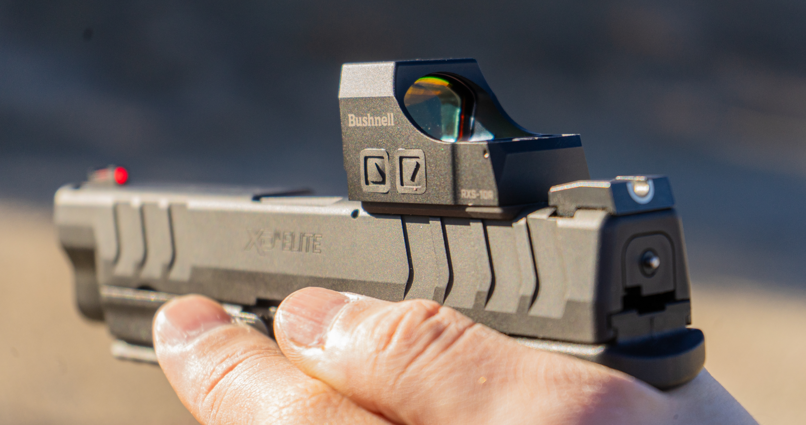 Hand holding a handgun with a red dot sight against a blurred background