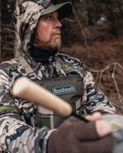 Person in camouflage gear with a Bushnell device, likely in a hunting or outdoor setting.
