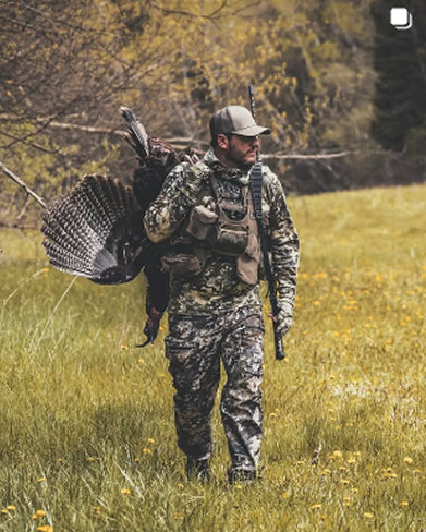 Hunter in camouflage walking through a field with a turkey on a shoulder strap.
