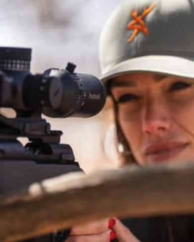 Person aiming a rifle with a scope, wearing a cap with a logo.