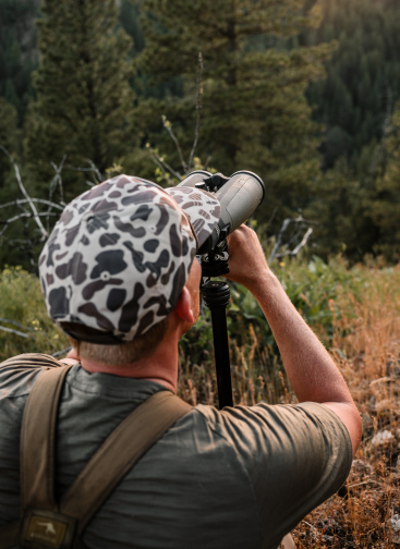 A man viewed from behind wearing a black and white animal print baseball cap and a green shirt, looking through a large binocular mounted on a tripod in a forest setting.