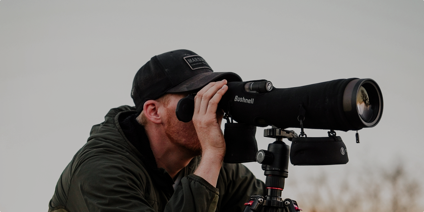 A person wearing a dark cap and jacket looks through a large spotting scope mounted on a tripod, focusing on a distant object outdoors against a light sky background.