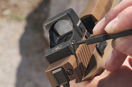 Person adjusting a rifle scope outdoors with rocks and grass in the background