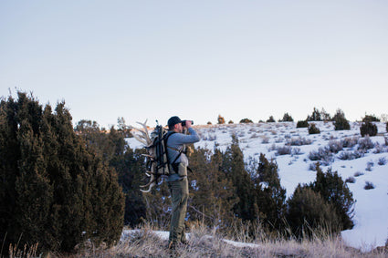 A person stands on a snowy hillside with green shrubs, carrying antlers on their backpack and looking through binoculars toward the horizon under a clear sky.