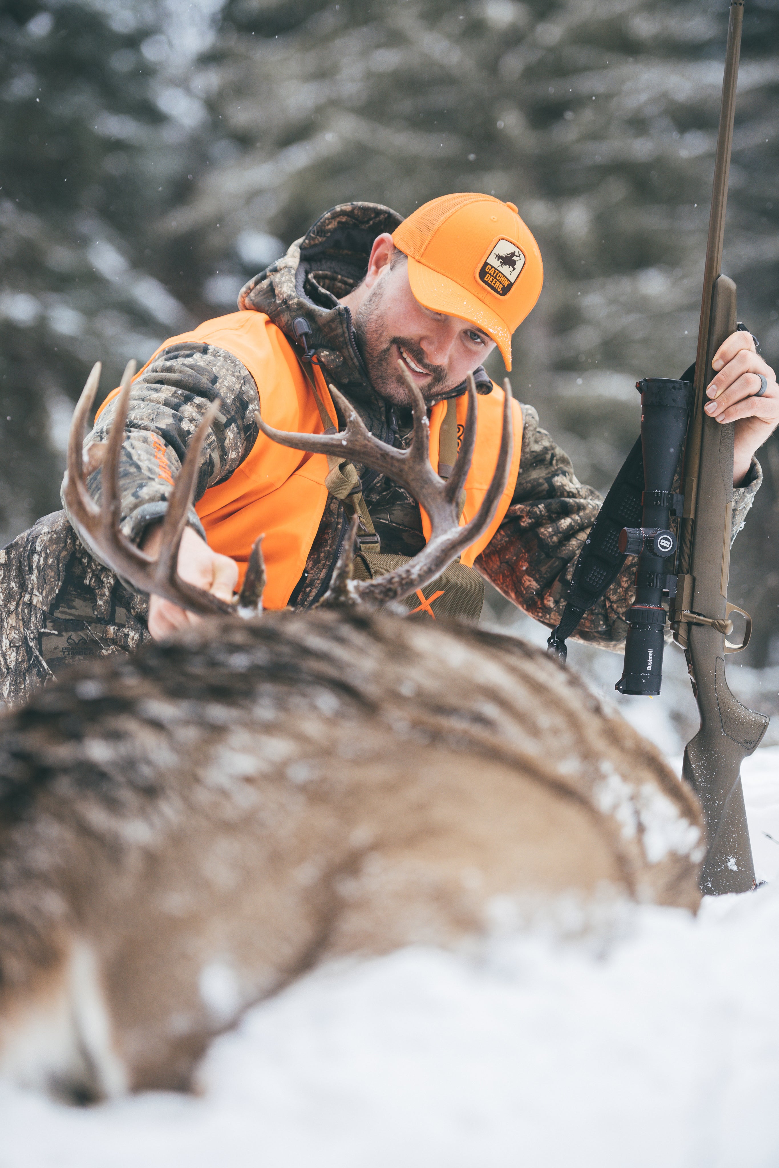 A smiling hunter in camouflage and an orange vest holds a rifle and touches the antlers of a large deer lying in the snow, surrounded by a snowy forest background.