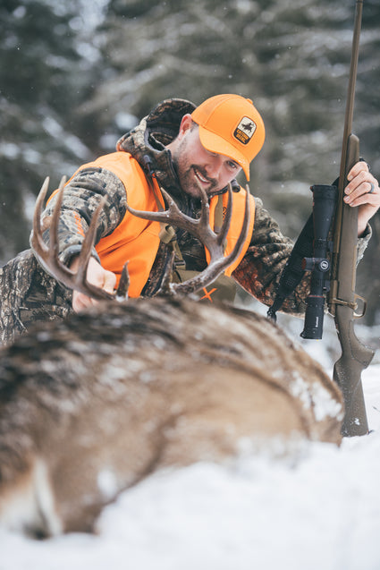 A smiling hunter in camouflage and an orange vest holds a rifle and touches the antlers of a large deer lying in the snow, surrounded by a snowy forest background.