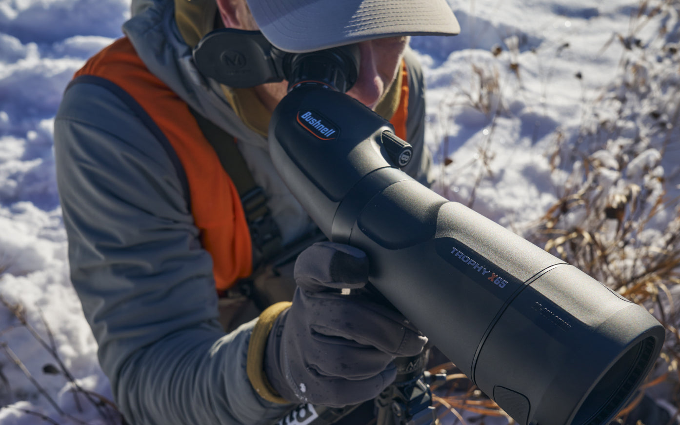A person in outdoor gear uses a spotting scope while kneeling in the snow, surrounded by dry grass. The scope is labeled TROPHY XLT and Bushnell.