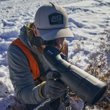 A person wearing a gray hat and orange vest is kneeling in the snow, looking through a large black spotting scope labeled “Trophy XLT.” Dry plants and snow are visible in the background.