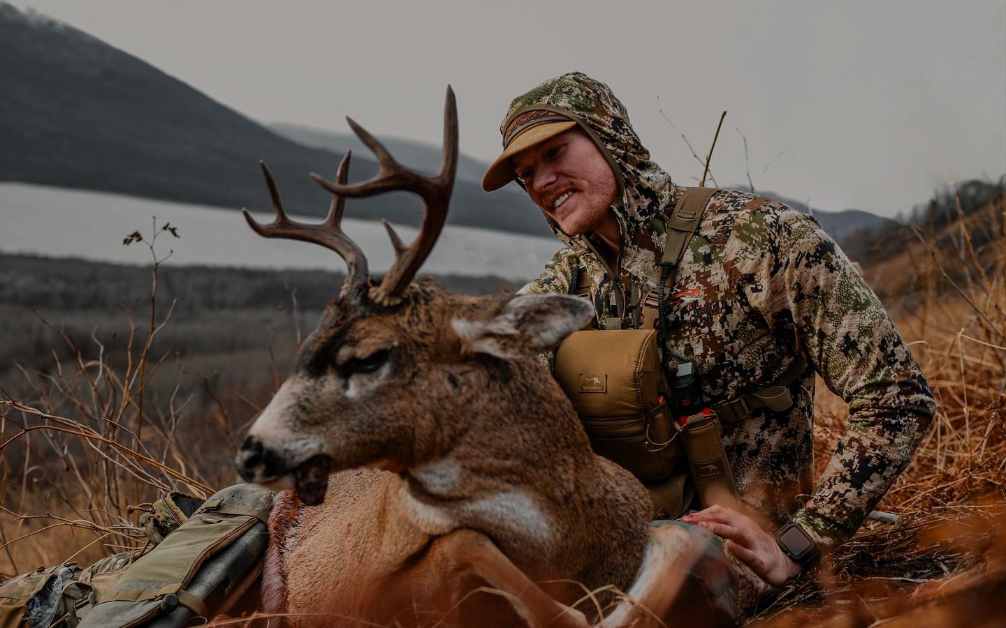 A man in camouflage clothing kneels on dry grass next to a large deer with antlers, smiling outdoors near a lake and mountains under a cloudy sky.