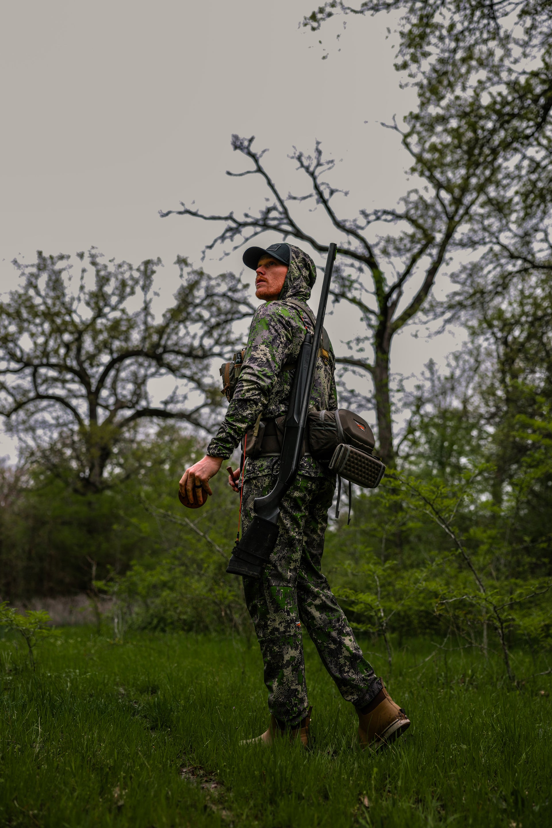 A person in camouflage clothing and boots stands in a grassy, wooded area holding a firearm and looking back over their shoulder; branches and trees fill the background under a cloudy sky.