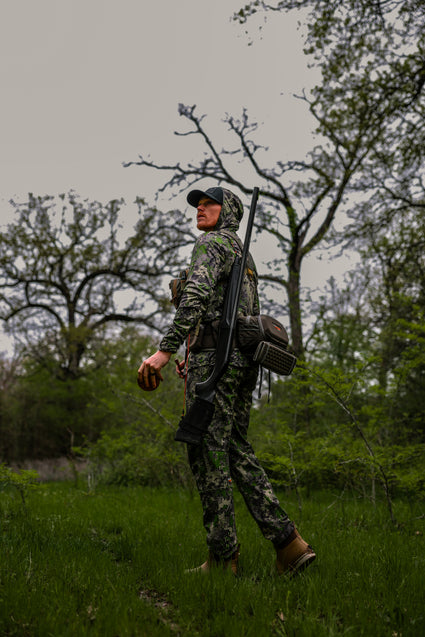 A person in camouflage clothing and boots stands in a grassy, wooded area holding a firearm and looking back over their shoulder; branches and trees fill the background under a cloudy sky.