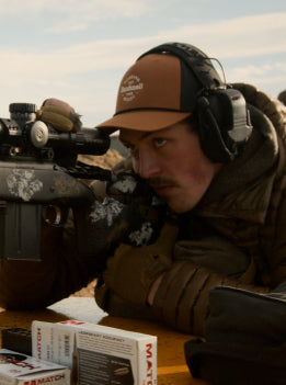 Man in hunting gear with rifle and binoculars, looking through scope.