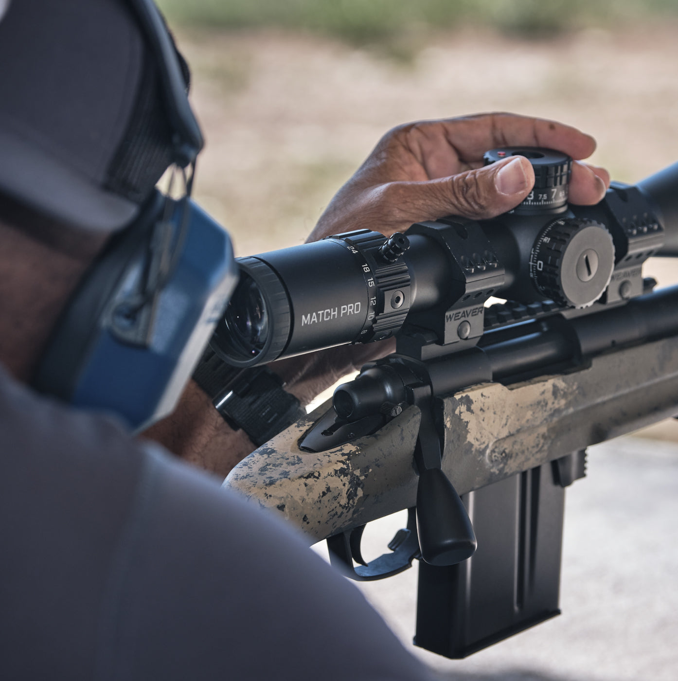Person holding a rifle with a scope, wearing ear protection, in an outdoor setting.