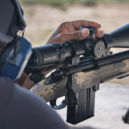 Person holding a rifle with a scope, wearing a helmet, in an outdoor setting.