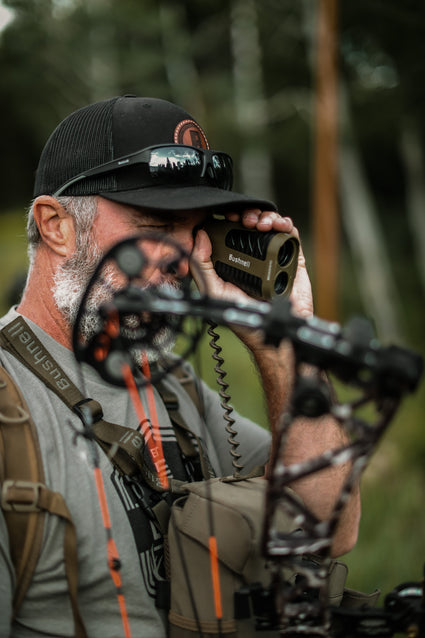 A bearded man wearing a cap uses a Bushnell rangefinder to measure distance while holding a compound bow outdoors, surrounded by blurred greenery.