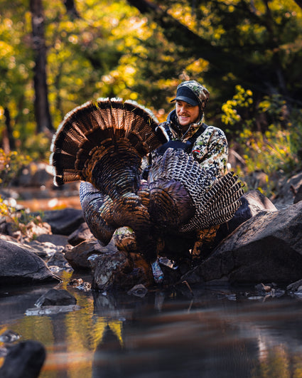 A person in camouflage clothing kneels by a stream, proudly holding a large wild turkey with vibrant feathers; trees with autumn foliage are in the background.