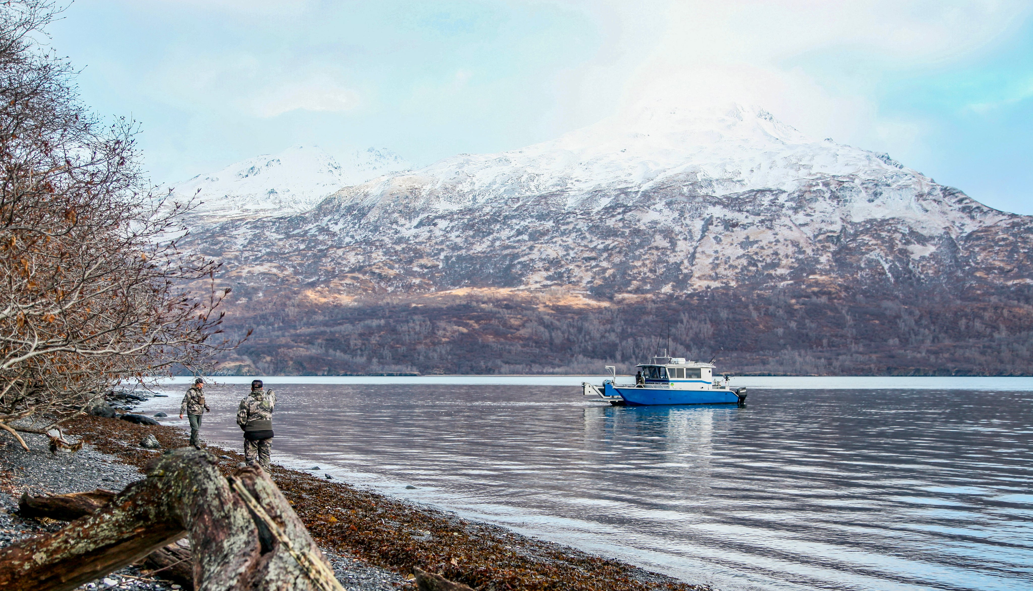 Two people stand on a rocky, tree-lined shore looking at a blue and white boat anchored on a calm lake, with snow-covered mountains in the background.