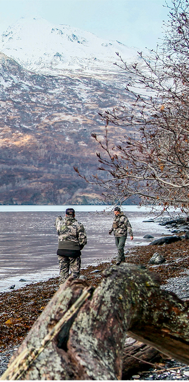 Two people in camouflage jackets stand on a rocky lakeshore with bare trees, facing a large, snow-covered mountain across the water. Fallen branches and autumn leaves cover the ground.