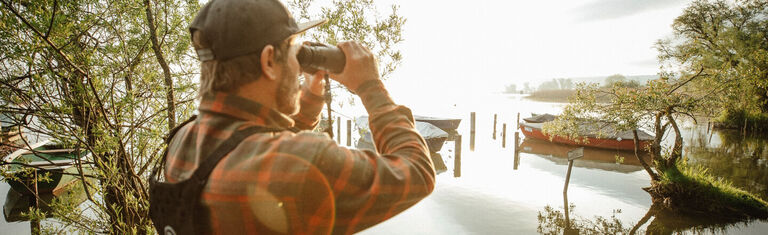 Man next to body of water looking through Bushnell Binoculars while using a Binocular Harness