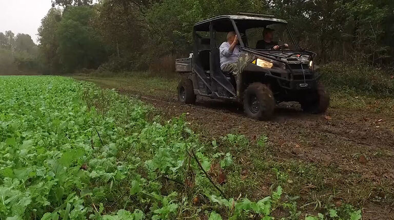 Two hunters in an ATV driving through a field next to wooded area