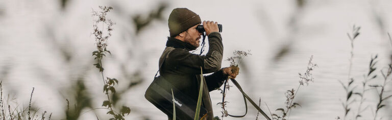 Man standing next to a body of water looking through a pair of Bushnell Binoculars